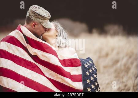 Shallow Focus Aufnahme eines amerikanischen Soldaten küsst seine liebevolle Frau, während sie in eine amerikanische Flagge gehüllt ist Stockfoto