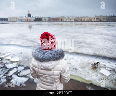 Frau im roten Hut in der Nähe von zugefrorenen Newa mit Ente und Isaak Kathedrale auf der anderen Seite im Winter Sankt-Petersburg, Russland Stockfoto