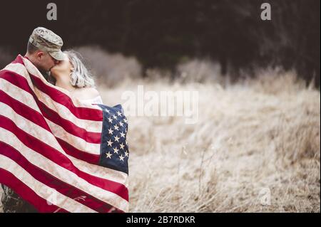 Shallow Focus Aufnahme eines amerikanischen Soldaten küsst seine liebevolle Frau, während sie in eine amerikanische Flagge gehüllt ist Stockfoto