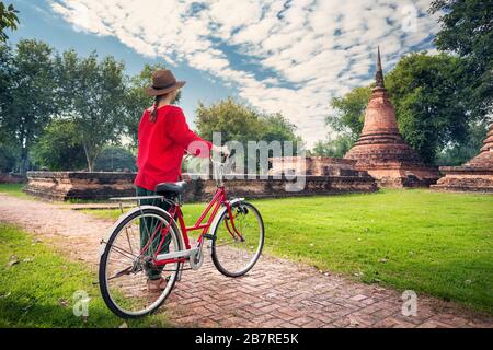 Frau im roten t-Shirt mit Vintage Fahrrad Blick auf alte ruiniert buddhistischer Tempel in erstaunliche Sukhothai Geschichtspark, Thailand Stockfoto