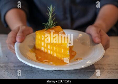 Nahaufnahme der Hände, die ein Stück Orangenkuchen auf Keramikplatte im Café halten und zeigen Stockfoto