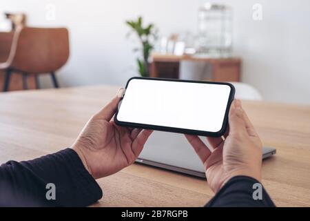 Verspottes Bild der Hände, die das schwarze Mobiltelefon mit einem leeren weißen Bildschirm horizontal halten, um es mit einem Laptop auf dem Tisch im Büro zu sehen Stockfoto