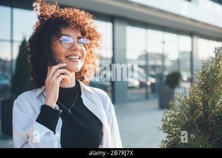 Der liebliche kaukasische Freiberufler mit lockigen Haaren hat ein Telefongespräch, während er draußen spaziert Stockfoto