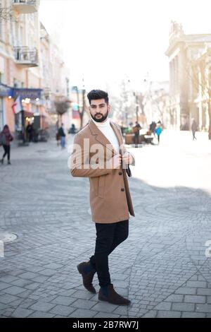 Porträt eines modisch gut gekleideten bärtigen Mannes auf der Straße. Junger indischer Mann in braunem Mantel, der sich im Freien aussehend darstellt. Stockfoto