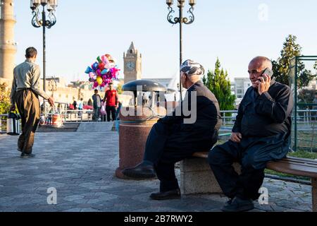 Irak, Iraq Kurdistan, Arbil, Erbil. Auf dem Park Shar sitzen zwei irakische kurdische Männer auf einer Bank. Eine befindet sich auf dem Telefon. OPN der linke Mann geht vorbei Stockfoto