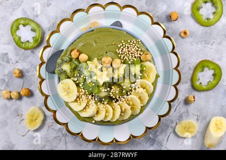 Smoothie-Schüssel aus grünem Obst mit sternförmigen Bananen, Kiwi, Haselnuss und gebucktem Quinoa-Korn auf grauem Grund Stockfoto