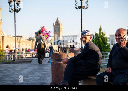 Irak, Iraq Kurdistan, Arbil, Erbil. Auf dem Park Shar sitzen zwei irakische kurdische Männer auf einer Bank. Eine befindet sich auf dem Telefon. OPN der linke Mann geht vorbei Stockfoto