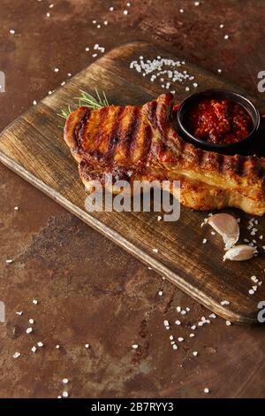 Hochwinkeliger Blick auf köstliches Steak mit Knoblauch und Tomatensauce auf Holzschneidebrett auf Steinoberfläche Stockfoto