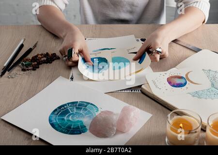 Beschnittener Blick auf den Astrologen, der Aquarellbilder mit Tierkreiszeichen auf Karten von Kerzen und Kristallen auf dem Tisch hält Stockfoto