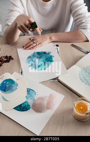 Beschnittener Blick auf den Astrologen, der Kristall über Wasserfarbenbildern mit Tierkreiszeichen auf Karten auf dem Tisch in der Kette hält Stockfoto