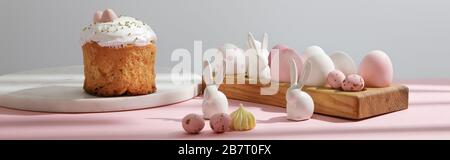 Ostereier auf Holzbrett mit dekorativen Kaninchen in der Nähe von osterkuchen auf rosa und grauem Hintergrund Stockfoto