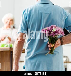 Selektive Fokussierung des Senioren-Mannes, der Blumenstrauß versteckt, während lächelnde Frau im Hintergrund kocht Stockfoto