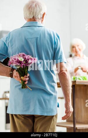 Selektive Fokussierung des Seniors, der Blumenstrauß versteckt, während lächelnde Frau in der Küche kocht Stockfoto