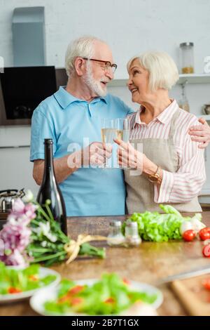 Selektive Fokussierung des Senioren-Paares, das sich beim Toasten mit Champagner durch Blumenstrauß und Gemüse auf Küchentisch anlächelt Stockfoto
