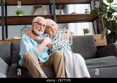 Niedriger Blickwinkel auf ältere Frauen, die Ehemann umarmen und zu Hause auf die Kamera auf der Couch schauen Stockfoto