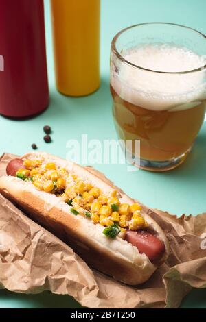 Nahaufnahme von heißem Hund mit Mais, Glas Bier und Flaschen mit Senf und Ketchup auf Blau Stockfoto