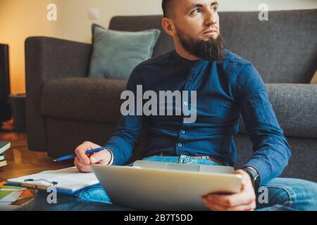 Bärtiger Geschäftsmann, der am Computer mit einigen Dokumenten auf dem Boden arbeitet und wegschaut, wenn er über etwas nachdenkt Stockfoto