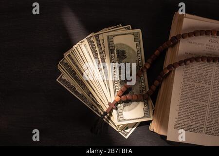 Draufsicht auf heilige bibel mit Geld und Rosary auf dunklem Hintergrund mit Sonnenlicht Stockfoto