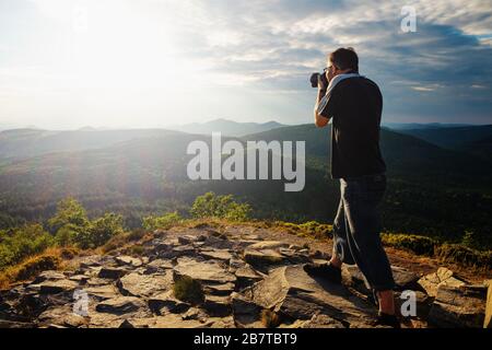 Mann drückt Kamera auf sein Gesicht und schaut in den Sucher. Touristen fotografieren in den Bergen Stockfoto