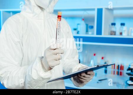 3/4-Ansicht des Wissenschaftlers im Haselmattenanzug mit Reagenzglas mit Coronavirus-Schriftzug Stockfoto