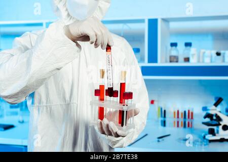 3/4-Ansicht des Wissenschaftlers in Hazmat-Anzug und Latexhandschuhen mit Reagenzglas mit Coronavirus-Schriftzug Stockfoto