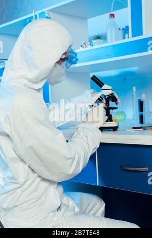 Wissenschaftler in Hazmattenanzug und Schutzbrille in der Nähe des Mikroskops im Labor Stockfoto