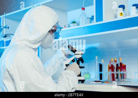 Wissenschaftler in Hazmattenanzug und Schutzbrille in der Nähe des Mikroskops Stockfoto