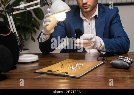 Beschnittener Blick auf den Schmuckgutachter, der den Ring mit Edelstein in der Schachtel neben der Lampe am Tisch hält Stockfoto