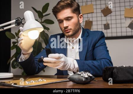 Gutaussehender Schmucksacheschätzer, der den Ring in der Schachtel in der Nähe von Lampe und Edelsteinen und Werkzeugen auf dem Tisch hält Stockfoto