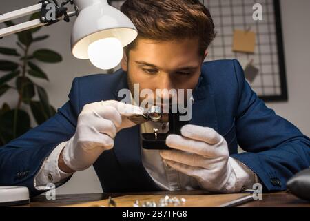 Gutaussehender Juwelierprüfer, der Ring mit Edelstein in der Box in der Nähe der Lampe auf dem Tisch untersucht Stockfoto