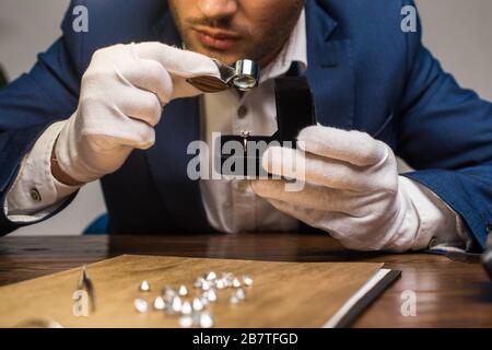 Beschnittenes Bild des Schmuckschätzers mit Lupe zur Untersuchung von Edelstein im Ring am Tisch auf grauem Hintergrund Stockfoto