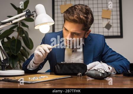 Gutaussehender Juwelenschätzer, der Edelstein in einer Zange in der Nähe von Schmuck auf dem Tisch in der Werkstatt untersucht Stockfoto