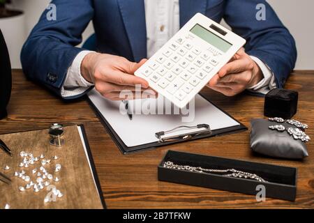 Beschnittener Blick auf den Schmucksachenschätzer, der den Rechner in der Nähe der Zwischenablage und Schmuck mit Schmucksteinen auf dem Tisch hält Stockfoto