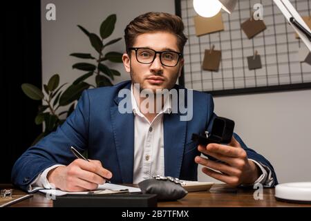 Schmucksacheschätzer, der die Kamera betrachtet, während er die Schachtel mit Ring hält und in der Nähe von Schmuck auf dem Tisch in der Werkstatt auf die Zwischenablage schreibt Stockfoto