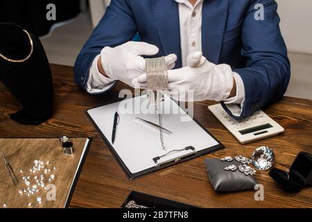 Beschnittener Blick auf Schmucksacheschätzer, der Halskette in der Nähe von Taschenrechner, Zwischenablage und Schmuck auf dem Tisch hält Stockfoto