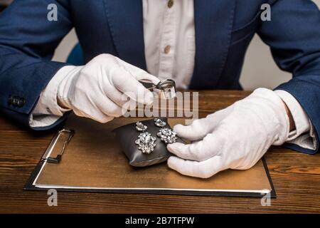 3/4-Ansicht des Juwelierschätzers, der Lupe und Einnahmen an Bord auf dem Tisch hält, isoliert auf Grau Stockfoto