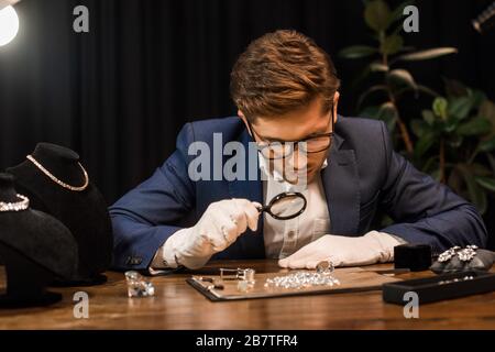 Gutaussehender Juwelierprüfer, der Edelsteine an Bord mit Lupe auf dem Tisch untersucht Stockfoto