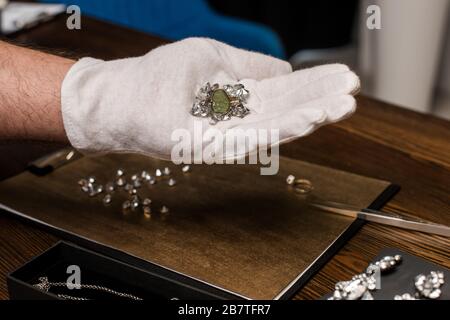 Beschnittener Blick auf den Juwelierschätzer, der Ring und Edelsteine in der Nähe von Schmuck und Brett auf dem Tisch hält Stockfoto