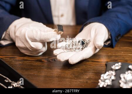 Beschnittener Blick auf den Schmucksachenschätzer, der Ring und Schmuck am Tisch hält, isoliert auf Schwarz Stockfoto