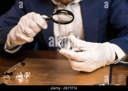 Beschnittener Blick auf den Schmucksachenschätzer, der Edelstein und Lupe in der Nähe von Schmuckringen an Bord auf einem Tisch hält, isoliert auf Schwarz Stockfoto