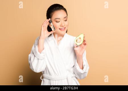 Fröhliche schöne asiatin im bademantel mit Avocado-Hälfte und Öl in Flasche auf beigefarbenem Hintergrund Stockfoto