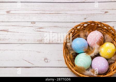 Draufsicht auf ostereier handbemalt in Pastellfarben und zarten Federn in einem Korbkorb auf Holztisch Stockfoto
