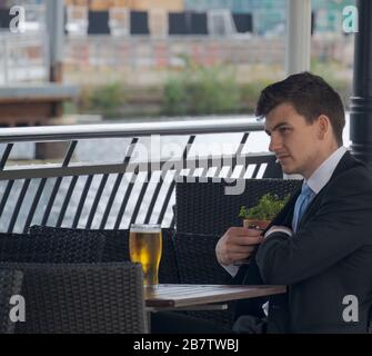 Der junge Geschäftsleute greift in seine Tasche, während er mit einem Glas Bier am Außentisch in Canary Wharf, London Docklands, Großbritannien sitzt. Stockfoto