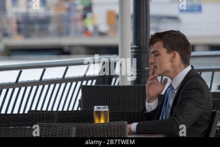 Der junge Geschäftsleute raucht eine Zigarette, während er mit einem Glas Bier am Außentisch in Canary Wharf, London Docklands, Großbritannien sitzt. Stockfoto