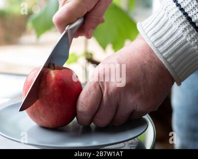 A bemannt die Hand mit einem Küchenmesser schneidet einen reifen roten apfel auf einer grauen Platte Stockfoto