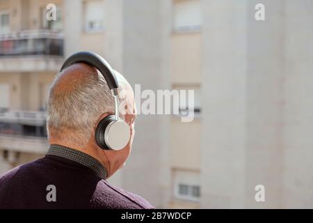 Nahaufnahme eines alten weißen Mann, von hinten gesehen, mit der drahtlose Kopfhörer im Freien, mit Blick auf eine Wohnung in einer Stadt Stockfoto