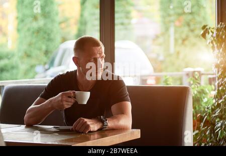 Gut aussehender sportlicher Mann hat eine Tasse Kaffee in einem Café, sieht weg, riesige Fenster mit Sonnenlicht hinter ihm auf dem Hintergrund Stockfoto