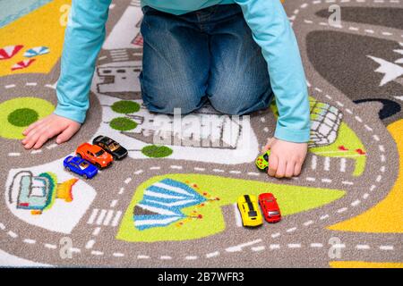 Fünf Jahre alter Junge, der auf einer Spielmatte mit Straßen Spielzeugautos spielt und auslegt. Der Junge ist in blaue Jeans gekleidet und hält einen Krankenwagen. Stockfoto