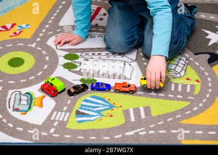 Fünf Jahre alter Junge, der auf einer Spielmatte mit Straßen Spielzeugautos spielt und auslegt. Die Autos haben lebhafte Farben und der Junge ist in blaue Jeans gekleidet. Stockfoto