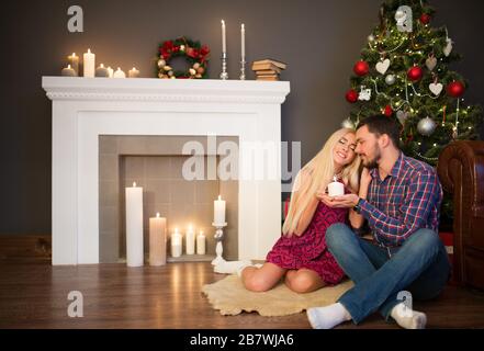 Schönes junges Paar charmantes langhaariges Mädchen und gutaussehender Flusspferd freuen sich am Heiligabend sitzend, Kamin mit einer Kerze in den Händen aga Stockfoto